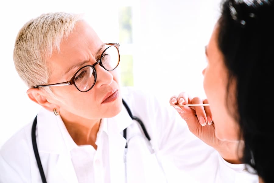 Caucasian mature senior female family doctor general practitioner checking examining attentively female patient`s throat with medicine stick